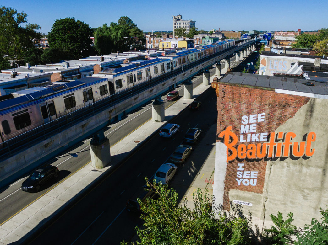 The elevated train runs by a painted mural that reads "See me like I see you: beautiful"