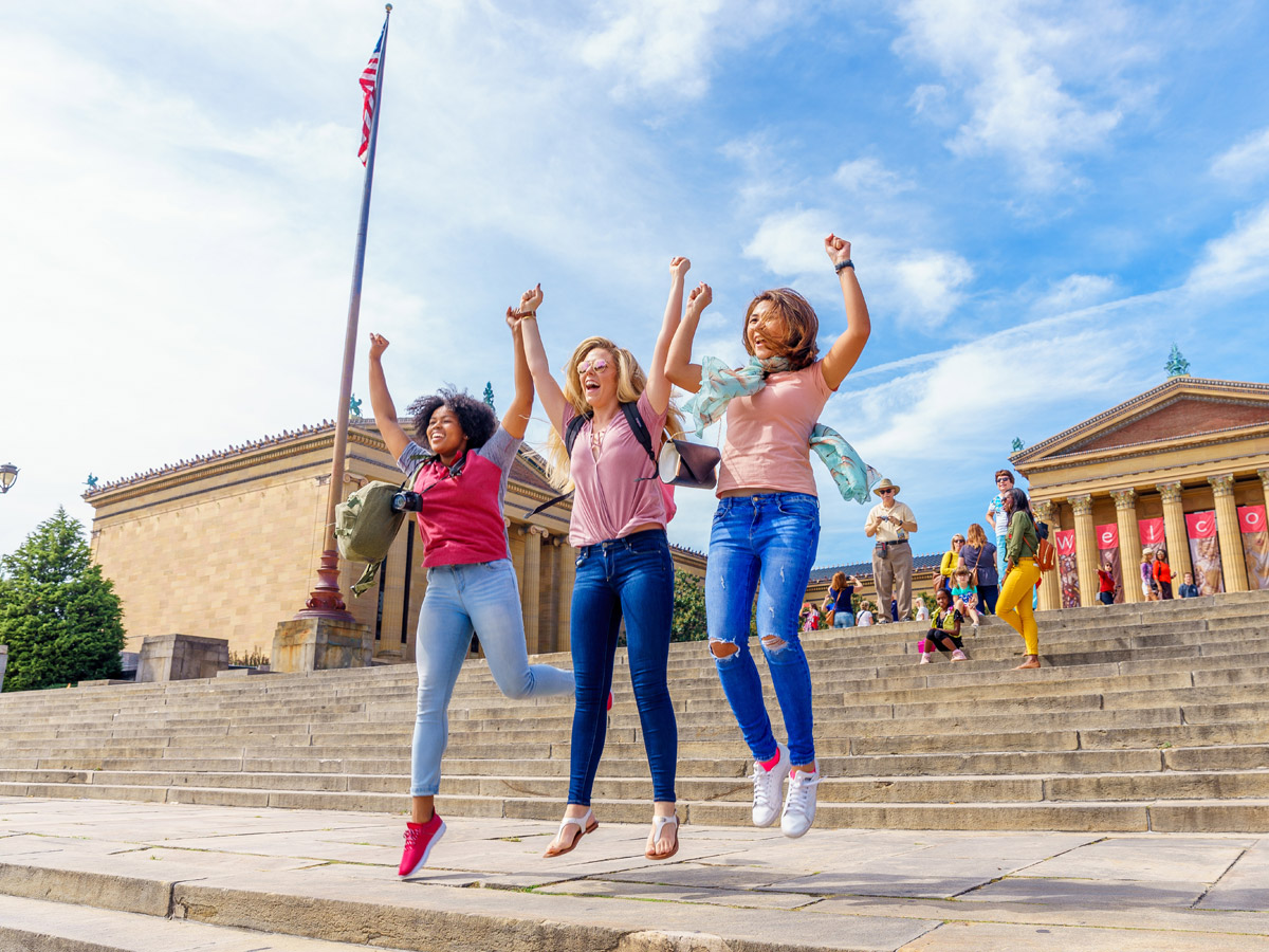 Three women jump in the air and hold their arms over their heads imitating Rocky on the steps of the Philadelphia Museum of Art