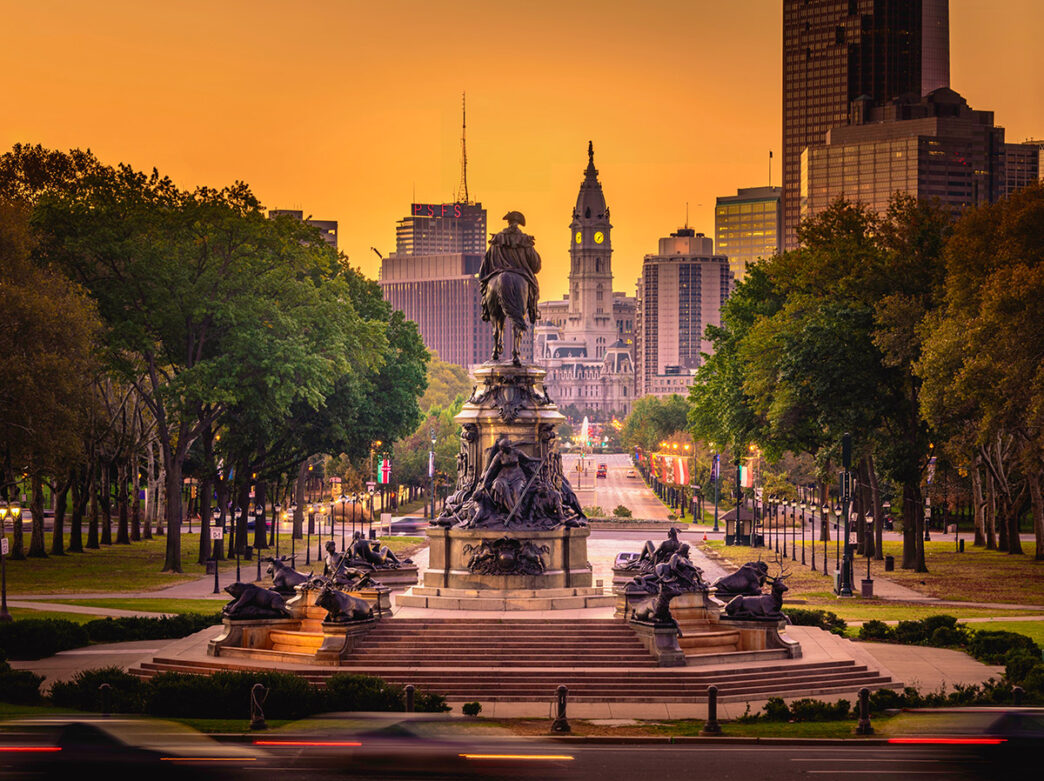 Against an orange sky, Philadelphia's Washington Monument statue on the Benjamin Franklin Parkway with City Hall in the background