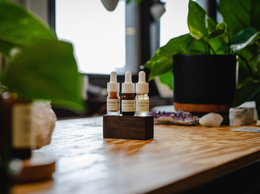 Three Sabbatical Beauty serums are displayed on a wooden table with green plants framing the photo.