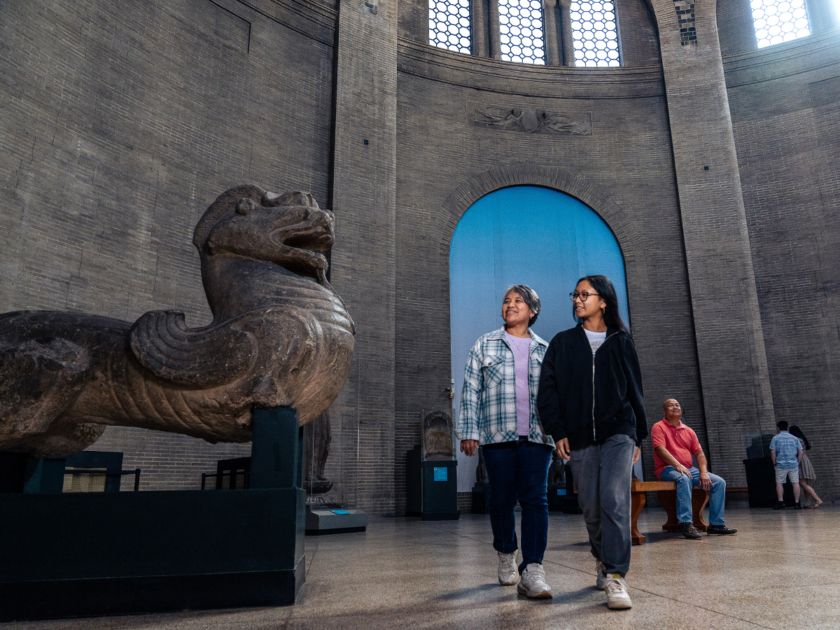 Two people walk past a large stone sculpture on display in the Asia Galleries rotunda at the Penn Museum.