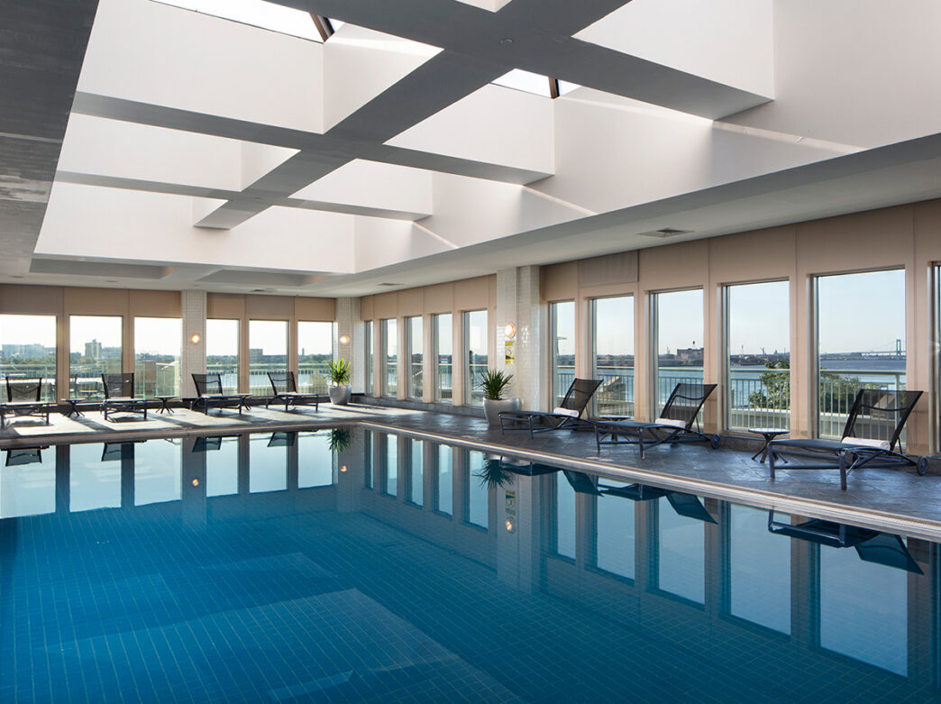 An indoor pool is filled with clear water and is surrounded by a pool deck with lounge chairs and floor to ceiling windows.