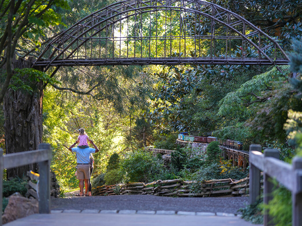 A father carries a child on their shoulders as they walk past the Garden Railway model trail at Morris Arboretum in Philadelphia.