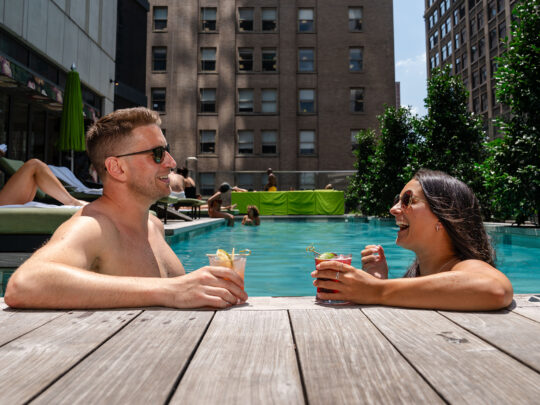 Two people relax in the pool at The W in Philadelphia. They both wear sunglasses and hold a drink in their hands.