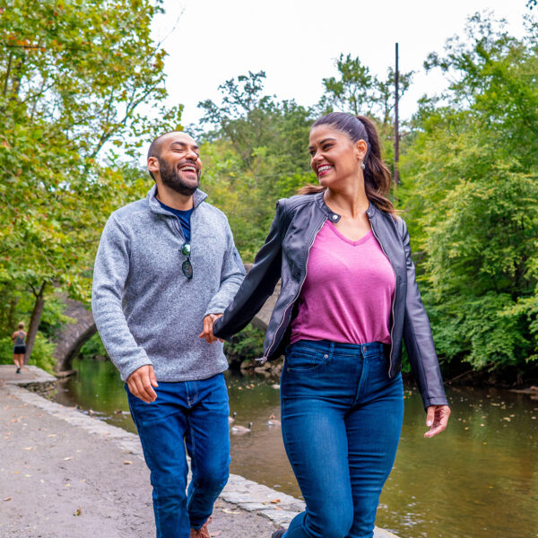 A woman wearing a pink shirt, leather jacket and jeans walks forward along a gravel trail while holding hands with a man behind her wearing a gray sweater and jeans.