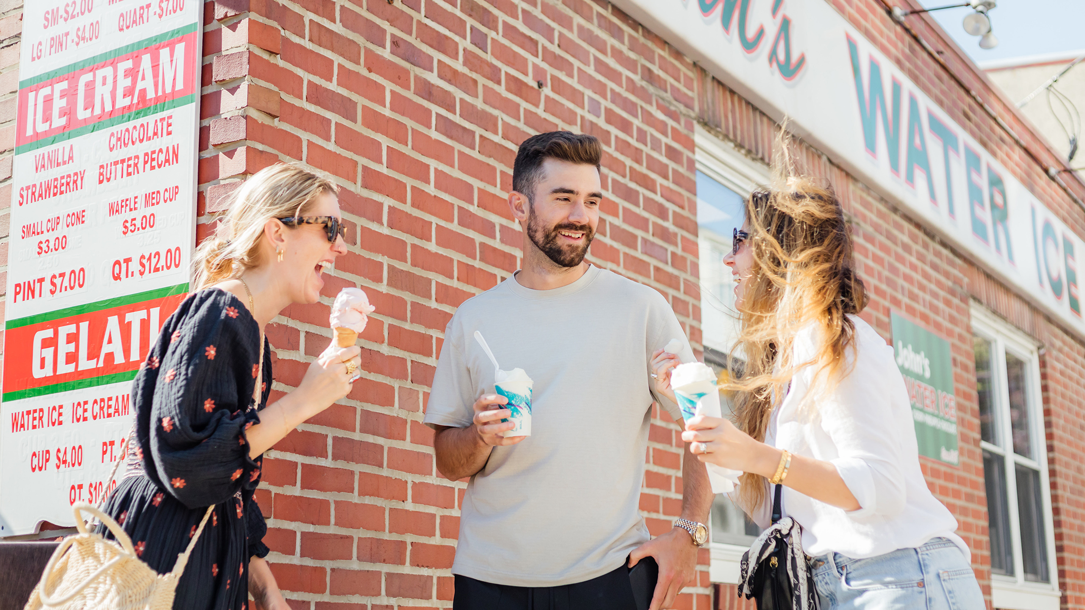 Three people stand in a group outside John's Water Ice laughing and eating water ice and ice cream.