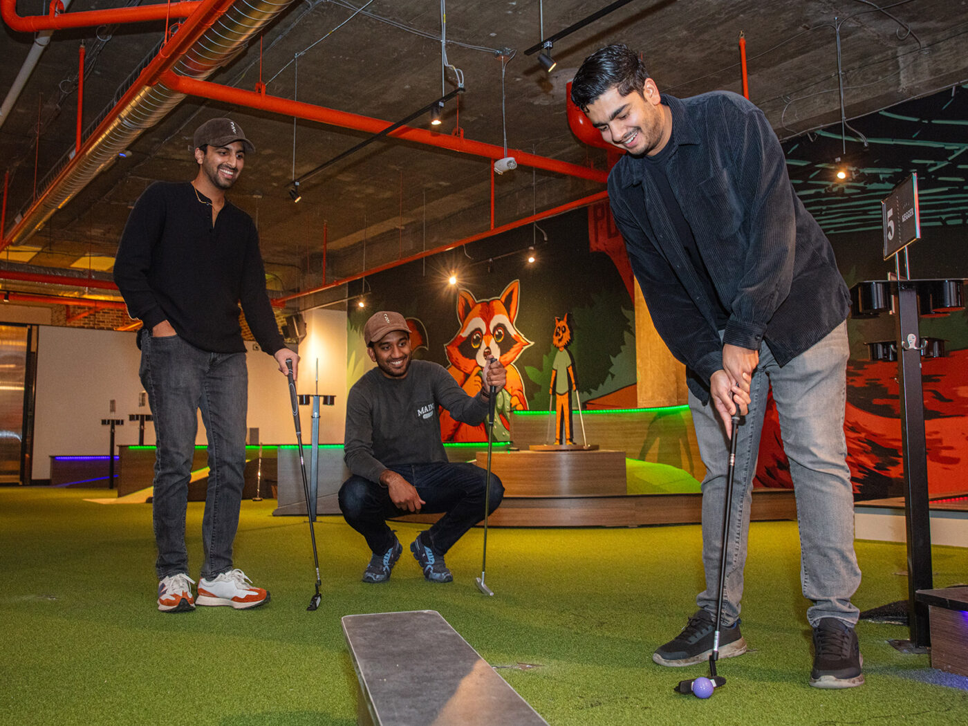 A group of young men dressed in casual black and gray outfits smile as they play a round of minigolf at Libertee Grounds.