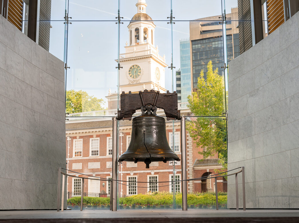 The bronze Liberty Bell is displayed in the Liberty Bell Center with floor to ceiling windows behind it. Through the glass windows, a brick building can be seen.