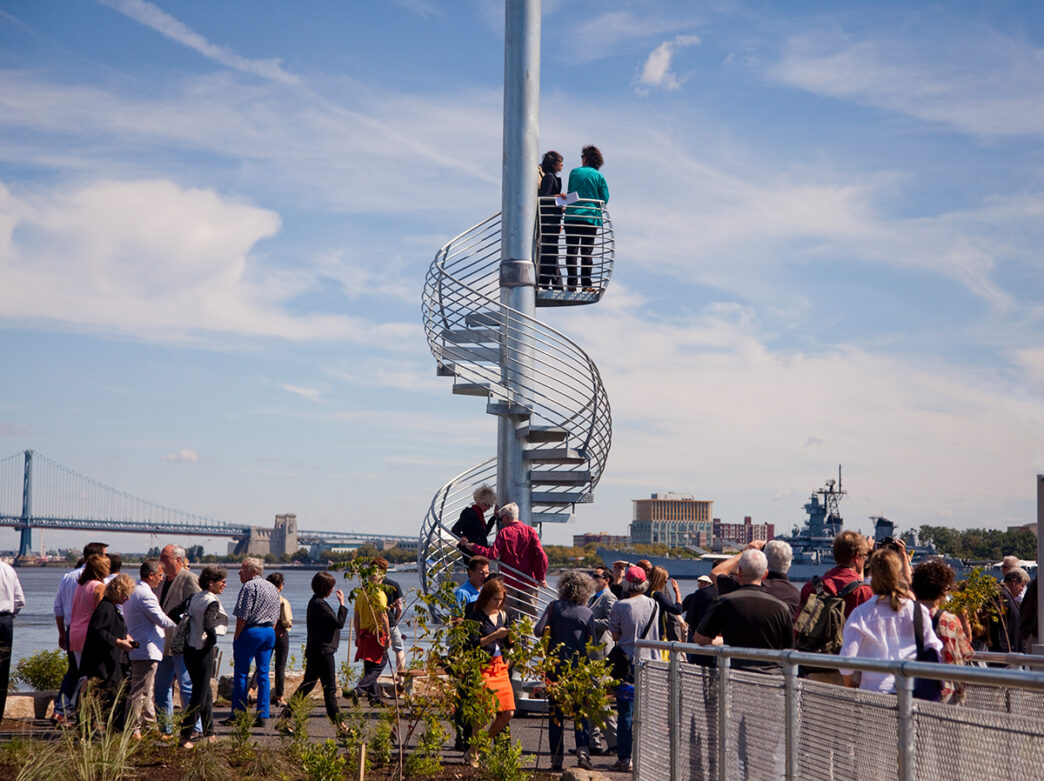 People stand at the top of metal spiral staircase looking out over the Delaware River. A crowd of people wait at the base of the staircase.