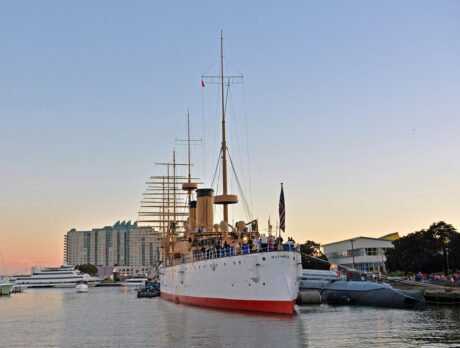 People stand on an old steel cruiser ship that is docked at the Delaware River Waterfront at sunset.
