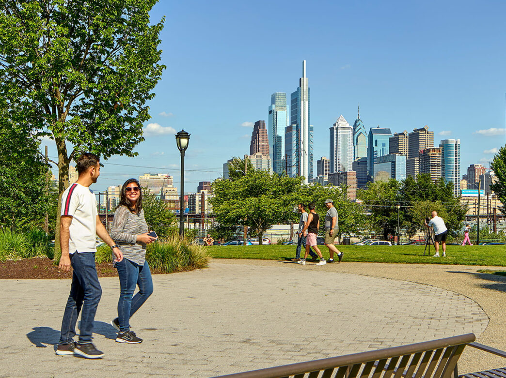 People walk along the pathways through Drexel Park with the Philadelphia skyline in the background.