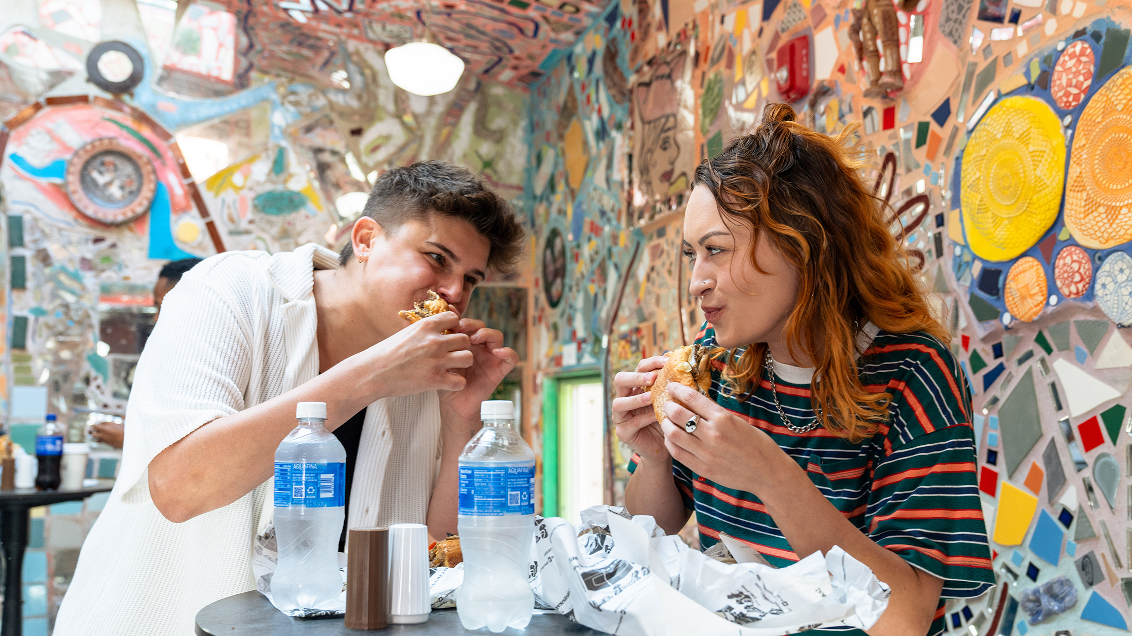 Two people stand at a table and eat cheesesteaks at Jim's Steaks on South Street. Behind them are colorful mosaics.