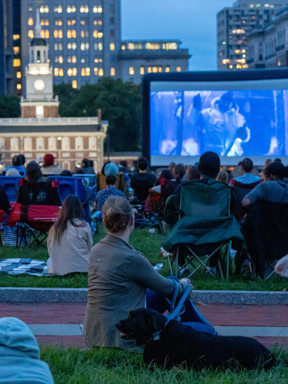People sit in camping chairs and on blankets with dogs in the grass on Independence Mall for Movies on the Mall. A large screen displays a movie. Independence Hall can be seen in the background.