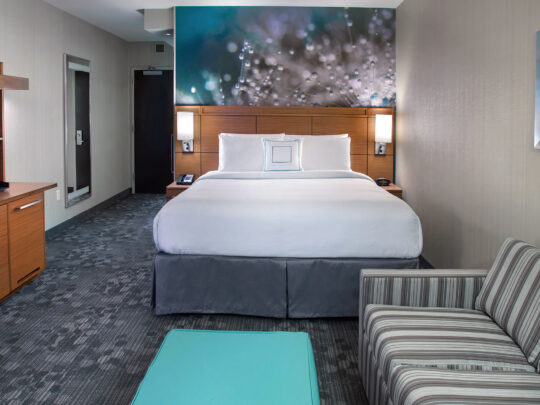 A hotel room inside the Courtyard South Philadelphia at the Navy Yard with a white and gray king-size bed, striped sofa, small teal table, and TV on a dresser.