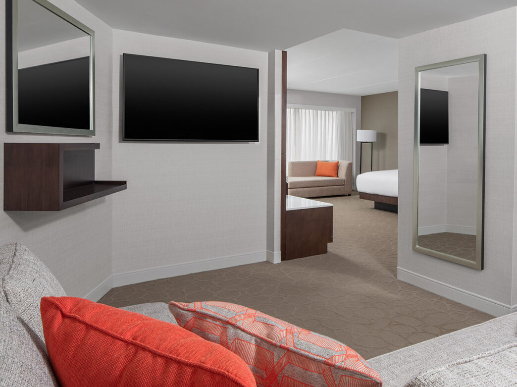 A view from a hotel suite's living room into the bedroom area, with a couch, bed, two mirrors and a television on the walls.