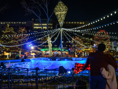 A couple stand with their arms around one another in front of a fountain decorated in colorful string lights at Electrical Spectacle in Franklin Square.