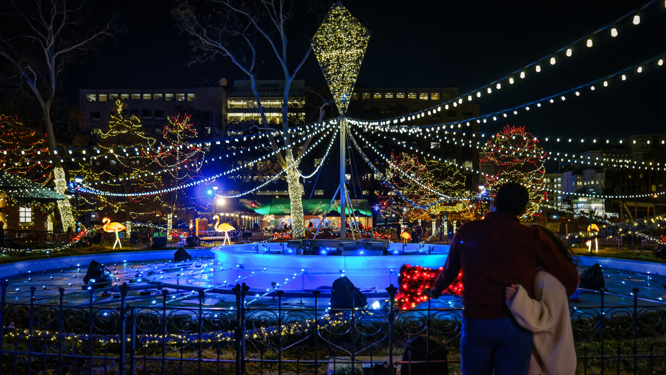 A couple stand with their arms around one another in front of a fountain decorated in colorful string lights at Electrical Spectacle in Franklin Square.