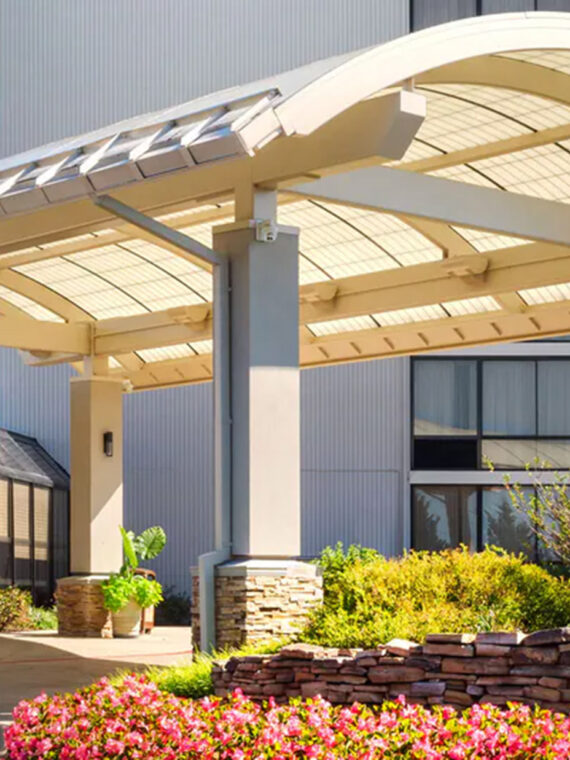 A welcoming hotel entrance featuring a modern arched canopy, a well-lit driveway, and attractive landscaping with colorful flowers and stone accents.