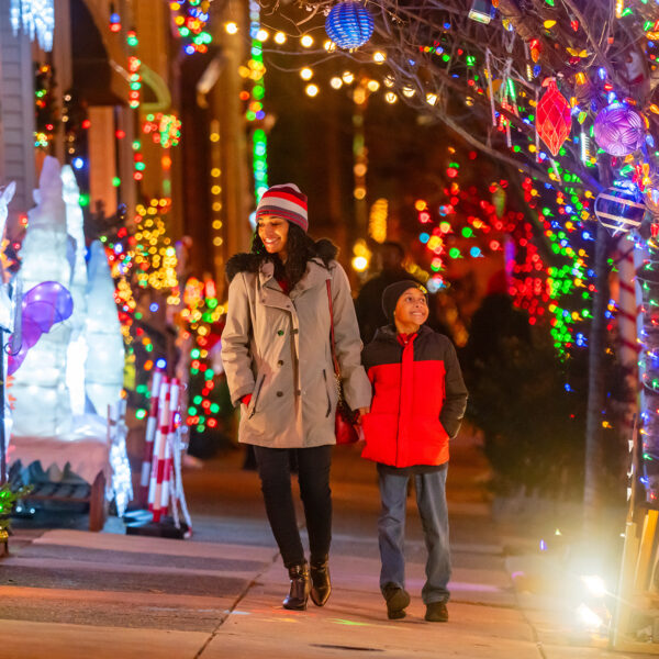 A mother and son hold hands and walk down the sidewalk at Miracle On 13th Street. They both smile and look at all of the colorful lights and decorations.
