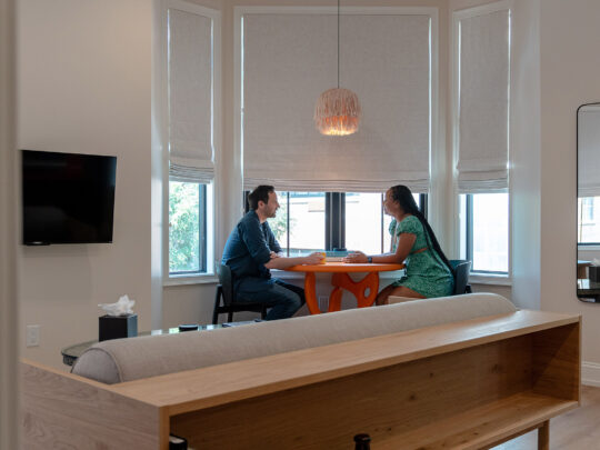 Surrounded by windows with the shades pulled halfway down, a couple sits at an orange table, under a tan fringe hanging light.