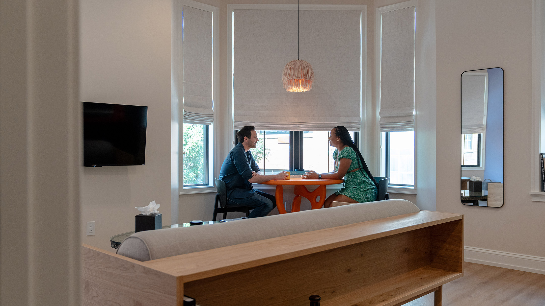 Surrounded by windows with the shades pulled halfway down, a couple sits at an orange table, under a tan fringe hanging light.