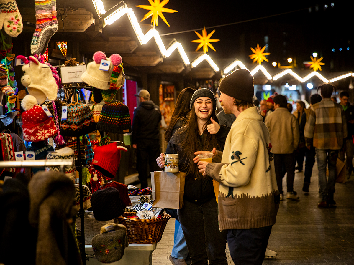 People hold pints of beer while shopping at the Christmas Village vendors. One of the people laughs happily.