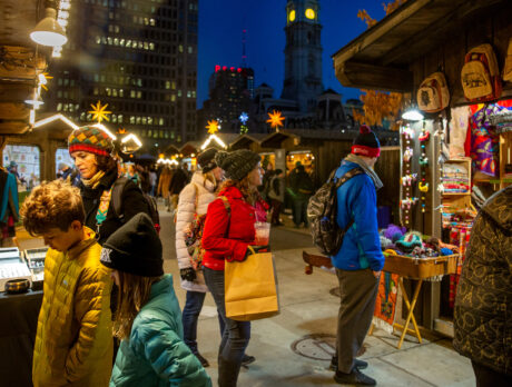People wearing jackets and hats shop at night at the Christmas Village in LOVE Park. The vendor huts have lights to illuminate their goods.