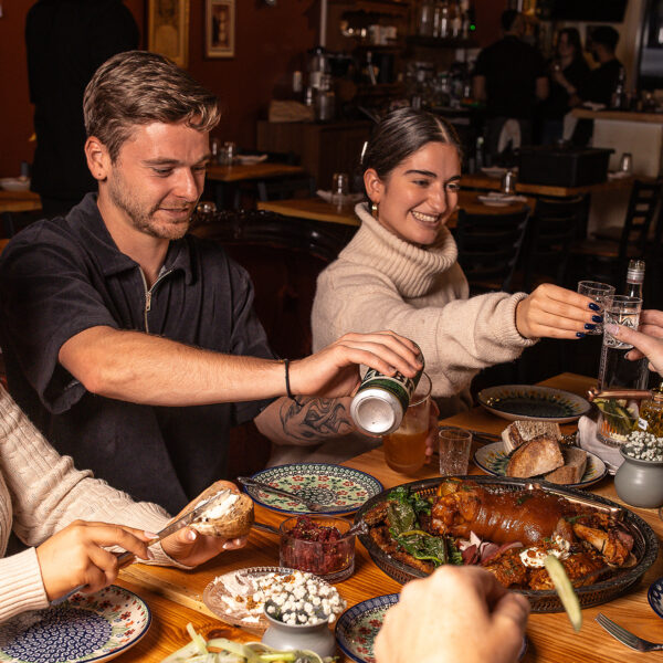 People sit at a dining table, toast glasses, pour beers and spread butter on bread at Little Walter's. Various food items are displayed on the table.
