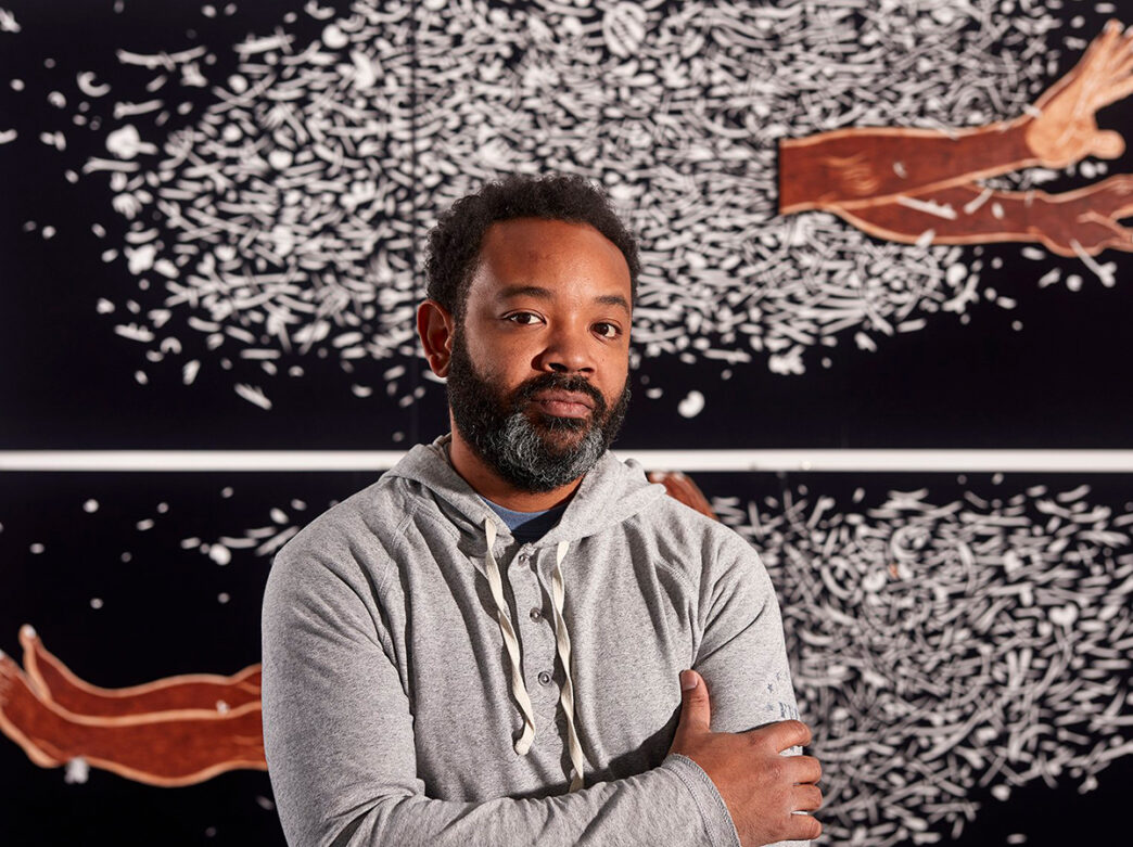 Artist William Villalongo wears a gray shirt and looks at the camera with his arms crossed. Behind him is a piece of white and black artwork.