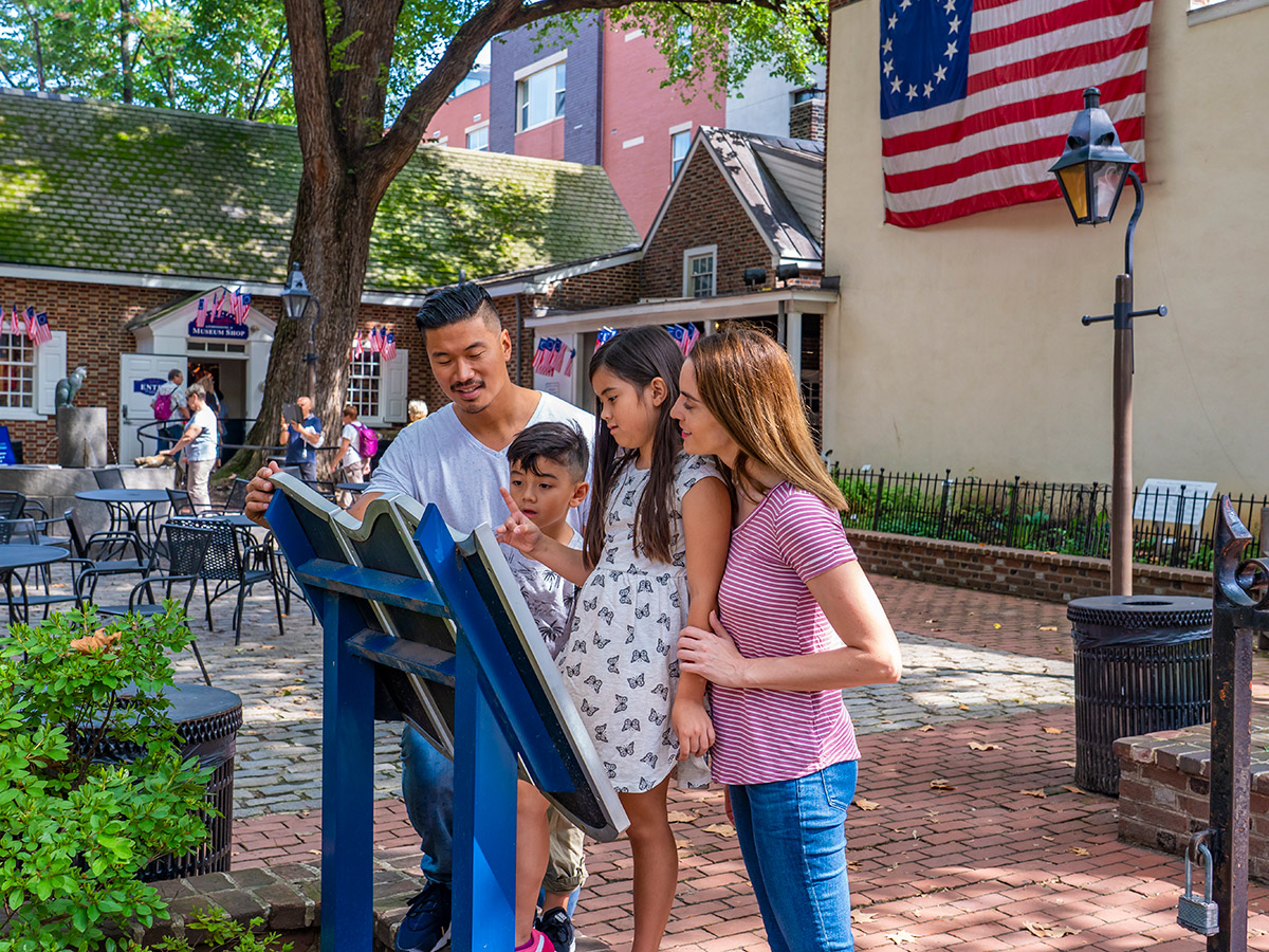 A casually dressed family stands in the courtyard of the Betsy Ross House and reads a plaque together.
