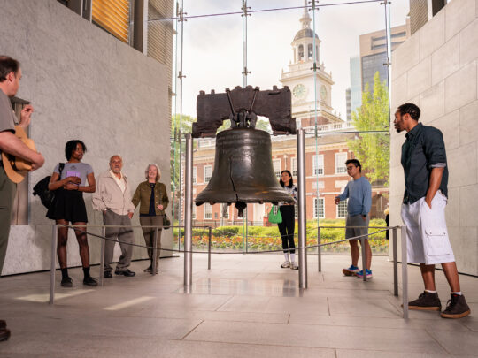 Seven people, including a National Park Service ranger, stand around and observe the Liberty Bell at Liberty Bell Center. Independence Hall is seen through the floor to ceiling windows behind the Liberty Bell.