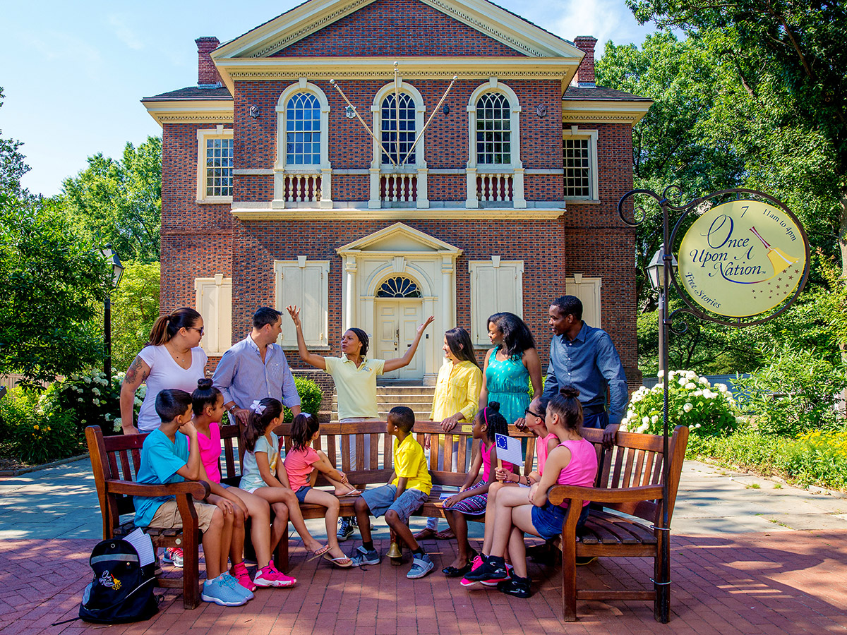 A storyteller, dressed in the yellow Once Upon a Nation uniform, tells a group of children and adults a story outside of Carpenters' Hall.