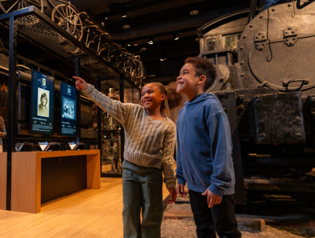One child points toward something while another child looks in that direction and smiles. Behind them is a steam locomotive in the Hamilton Collections Gallery at The Franklin Institute.