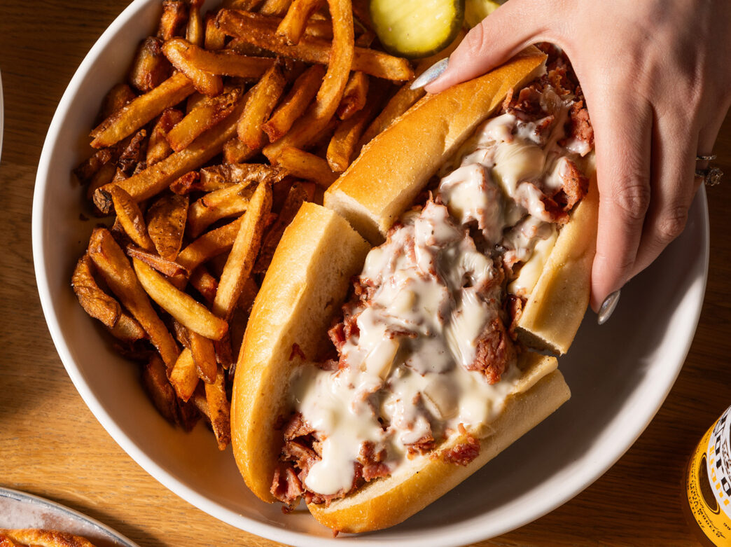 A hand with silver painted nails reaches for half a a cheesesteak on a round plate with crispy French fries and pickle slices. The Wit or Witowski Kielbasa Cheesesteak includes chipped kielbasa with onions and melted American cheese on a long roll.