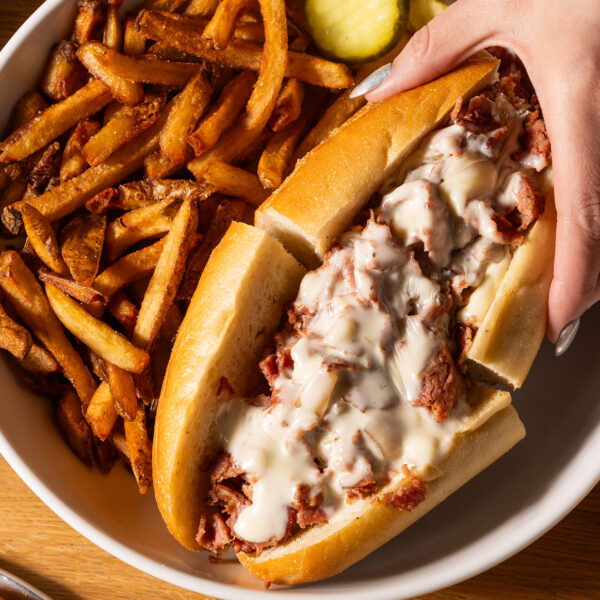 A hand with silver painted nails reaches for half a a cheesesteak on a round plate with crispy French fries and pickle slices. The Wit or Witowski Kielbasa Cheesesteak includes chipped kielbasa with onions and melted American cheese on a long roll.