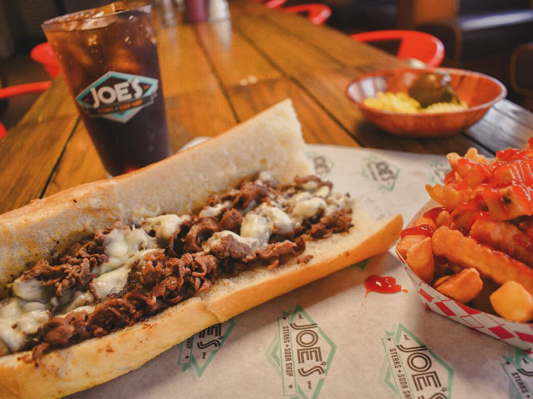 A cheesesteak with melted cheese is displayed on a tabletop with a side of fries and a soda.