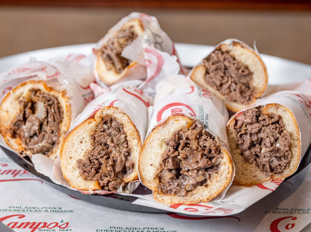 Six cheesesteaks wrapped in paper lay on a platter.