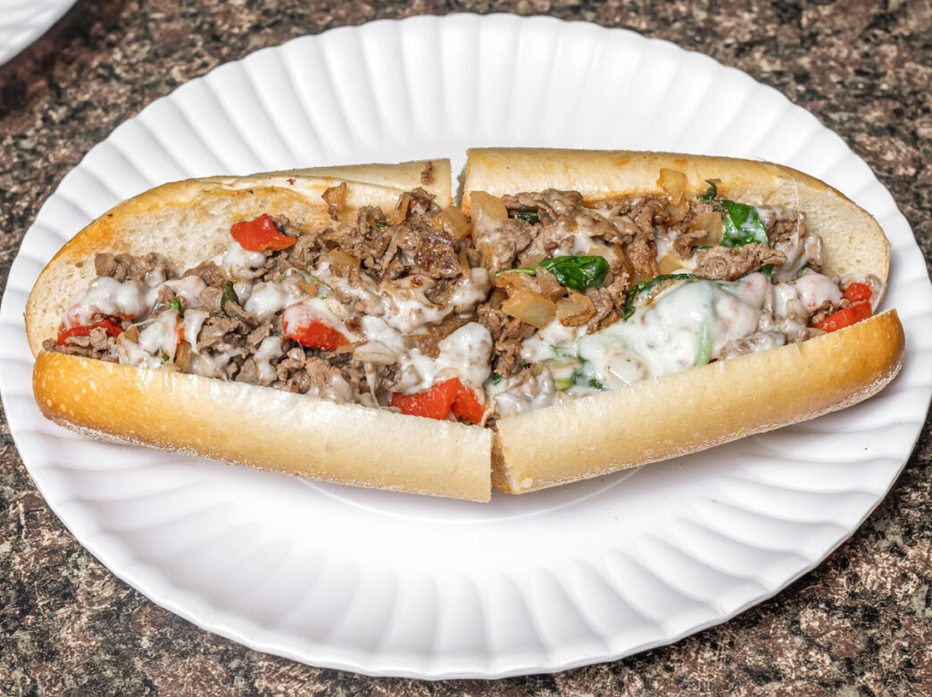 A beef cheesesteak topped with roasted peppers, spinach and provolone cheese is cut in half and displayed on a white plate at Ishkabibble's.