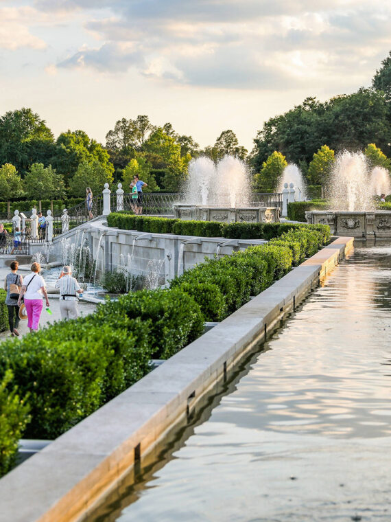 People walk through the Main Fountain Garden at Longwood Gardens. Green shrubs line the pathways between the spraying fountains.
