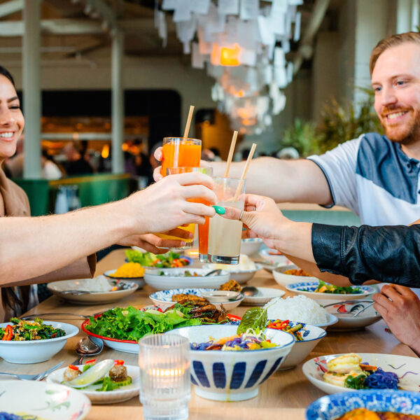 A group of smiling friends raise their glasses for a toast over a long table filled with colorful Thai cuisine.