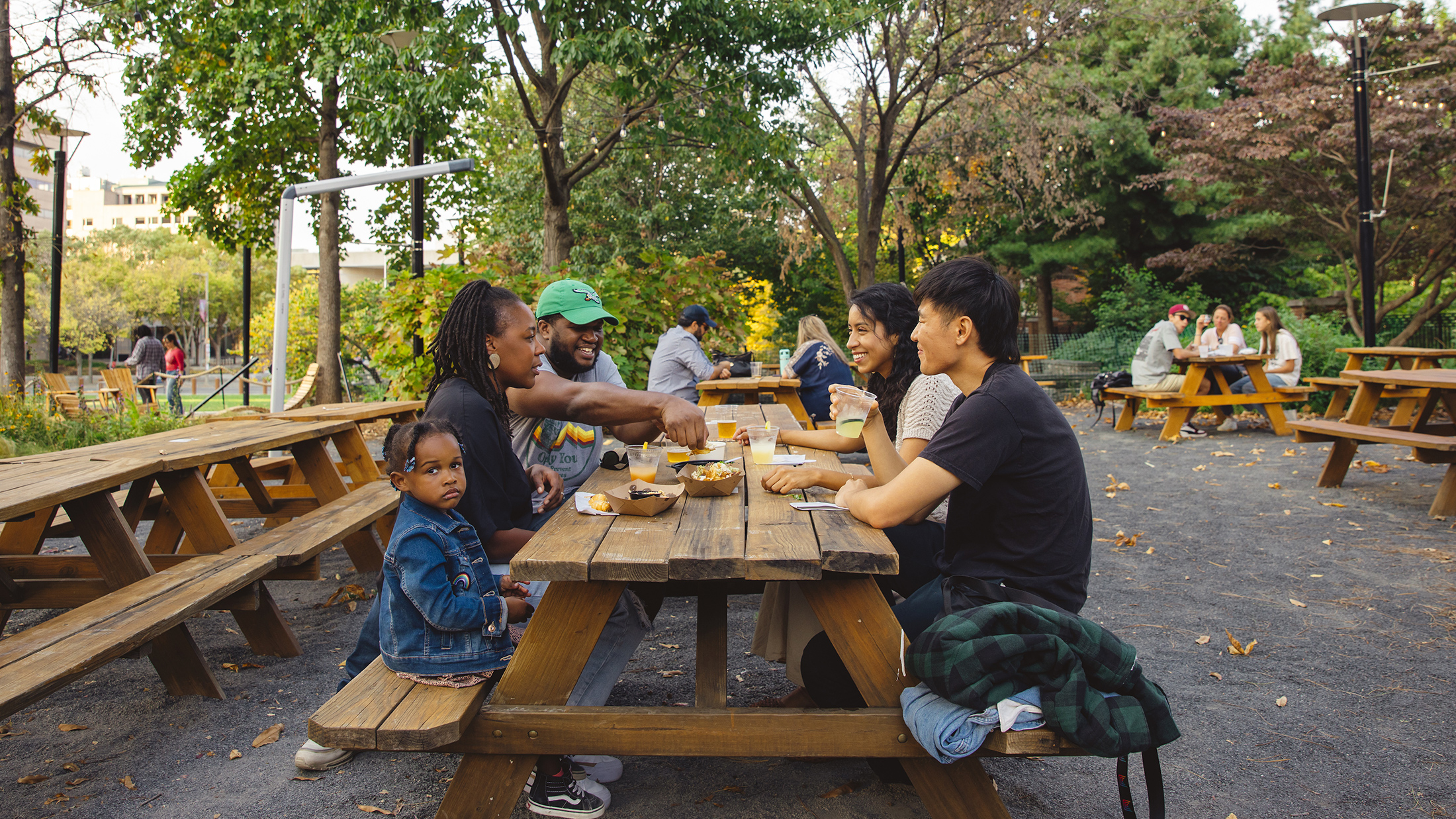 Four adults and one child sit at a picnic table and laugh while enjoying drinks and food items at Liberty Garden.
