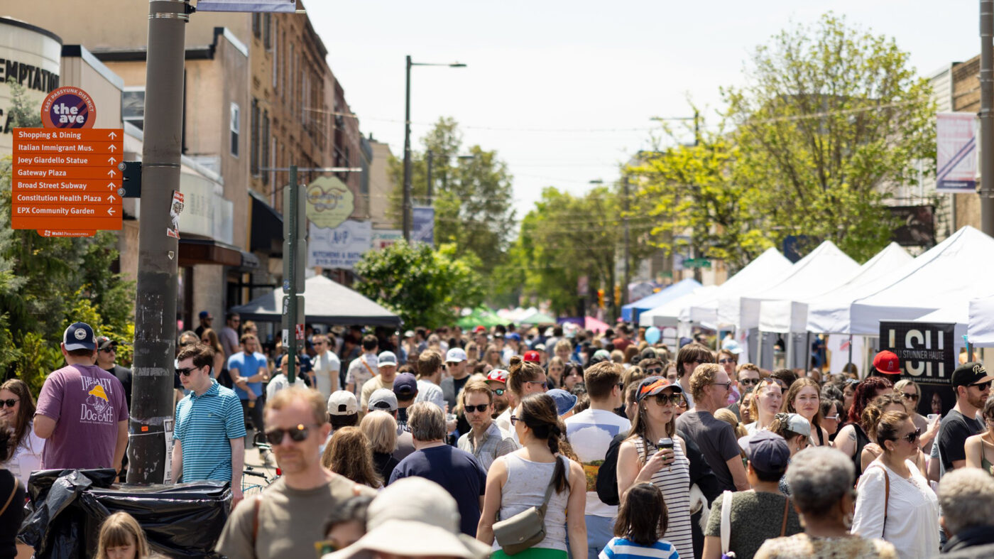 Crowds of people walk down East Passyunk Avenue during Flavors on the Avenue. The street is lined with white vender tents.