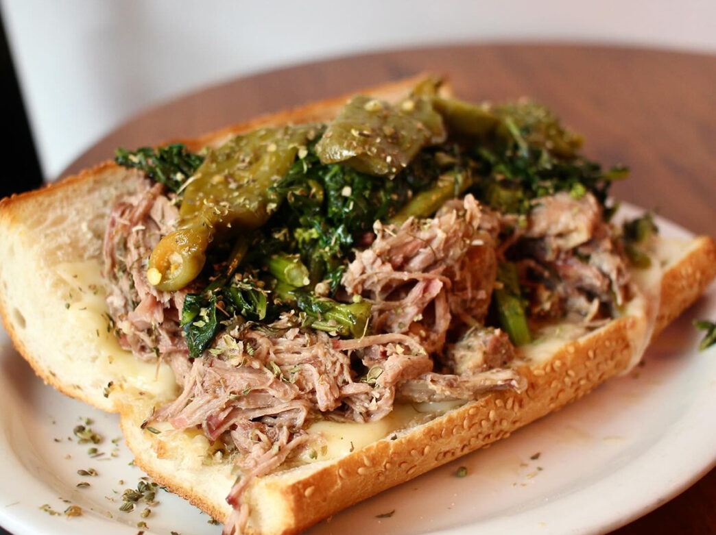 A close-up of the Arista sandwich from Paesano’s Philly Style. A seeded roll is filled with roasted pork, provolone, broccoli rabe and hot peppers.