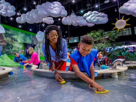 A smiling child and adult play with toy boats in an indoor water table at the Please Touch Museum. Hanging from the ceiling are fluffy clouds and a sun.