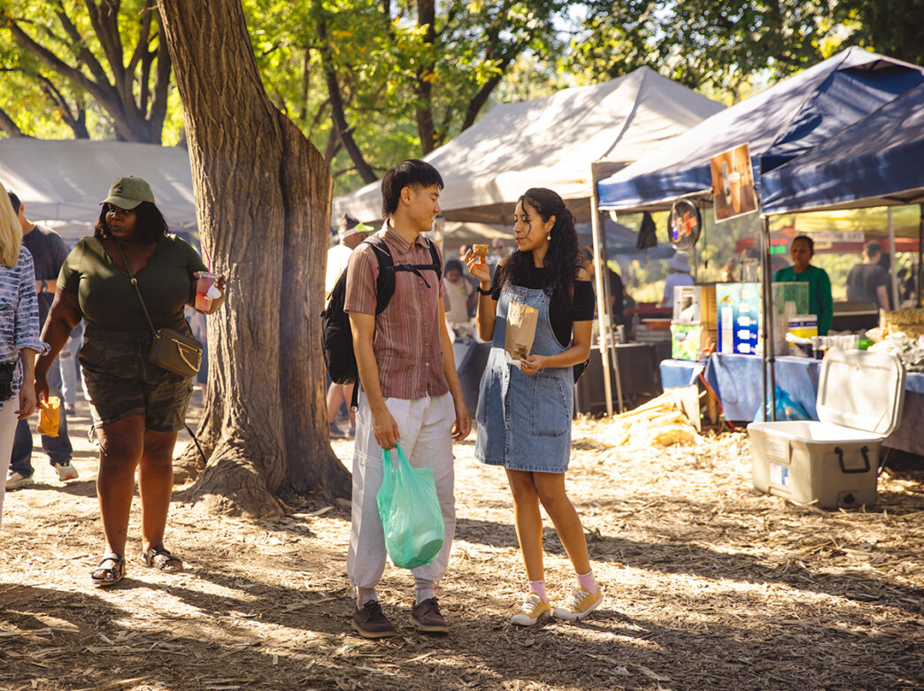 Two people walk through the Southeast Asian Market in FDR Park on a sunny day, holding snacks and a shopping bag, surrounded by vendor tents.