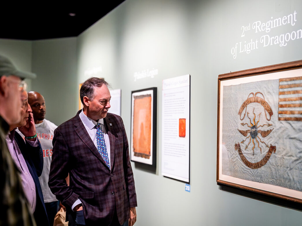 Visitors examine a framed historic flag on display at the Museum of the American Revolution's Banners of Liberty exhibit. The flag is displayed on a light green wall with interpretive text.