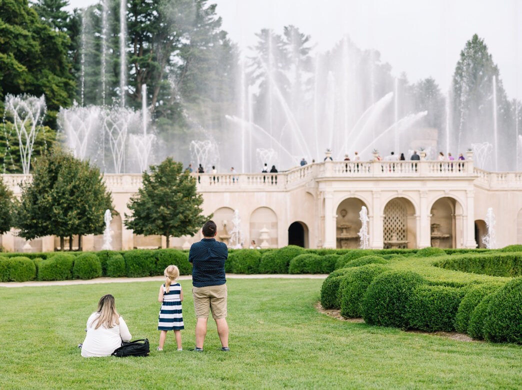 A family sits in the grass while looking up at a large fountain spraying water into the air at Longwood Gardens.