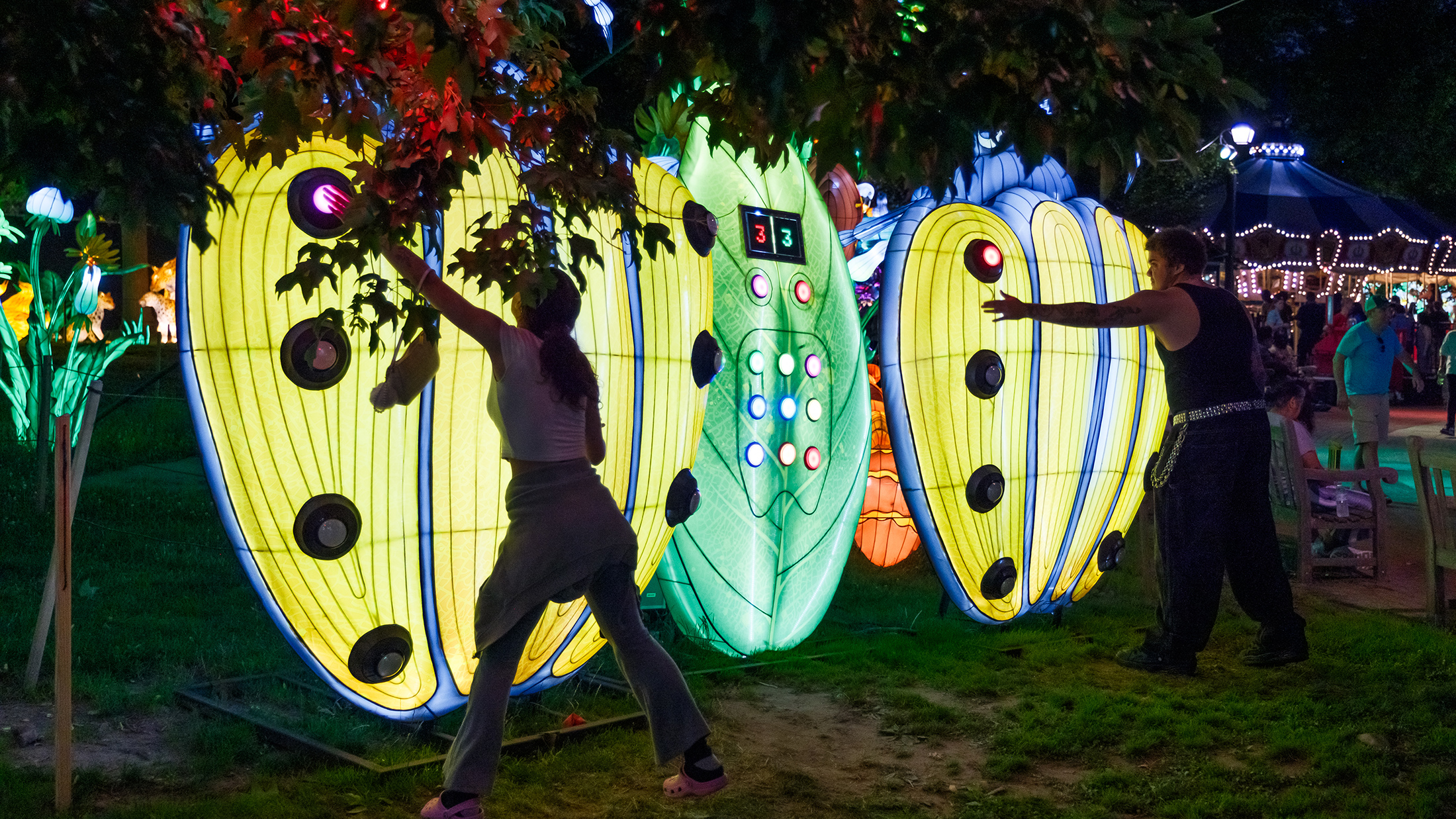 Two people play an illuminated, interactive game shaped like giant yellow beetles. The people hit light up buttons on the beetle wins.