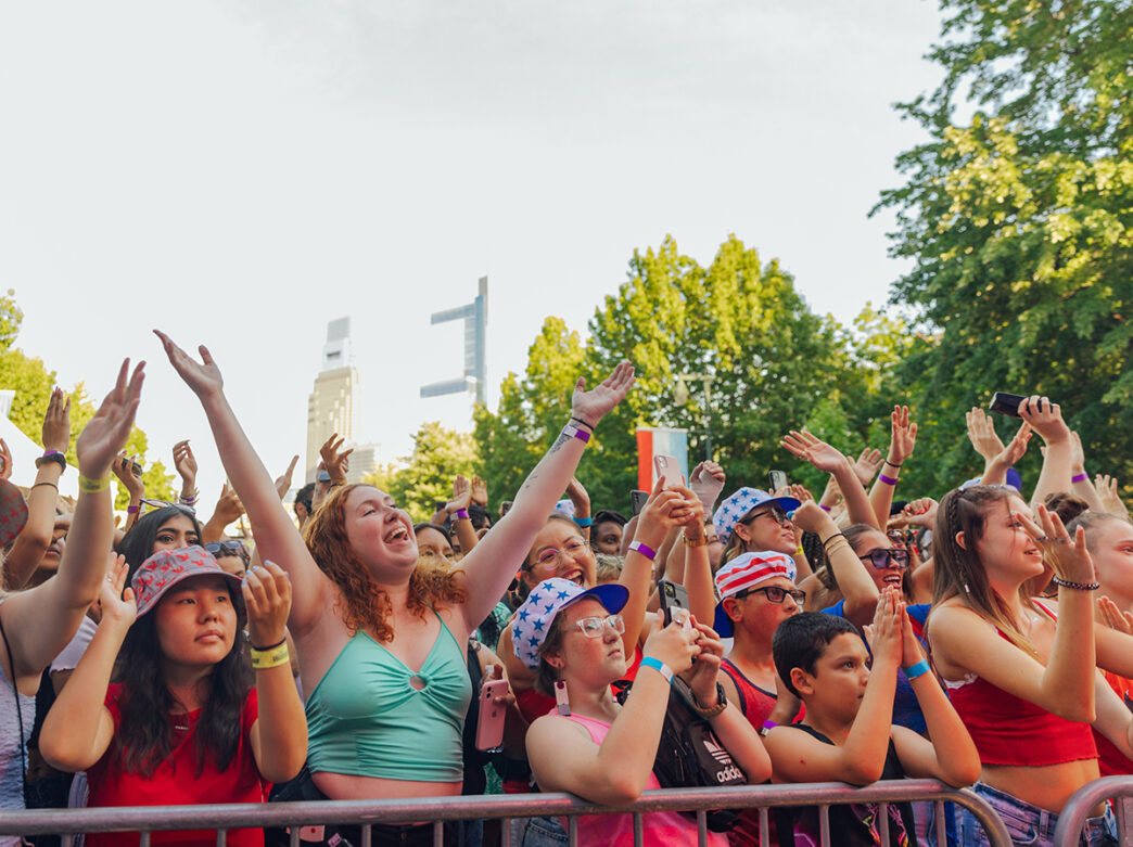 A lively crowd of people stand behind a metal railing, cheer and raise their hands in excitement while attending the July 4th concert in Philadelphia.