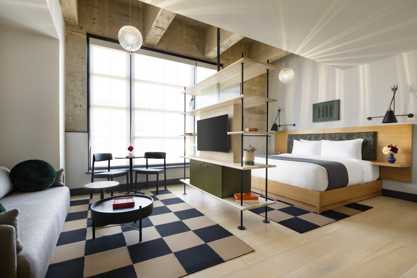 A bright, modern hotel room with a king bed, minimalist furniture, a checkerboard rug and central shelving unit with a mounted TV dividing the space.